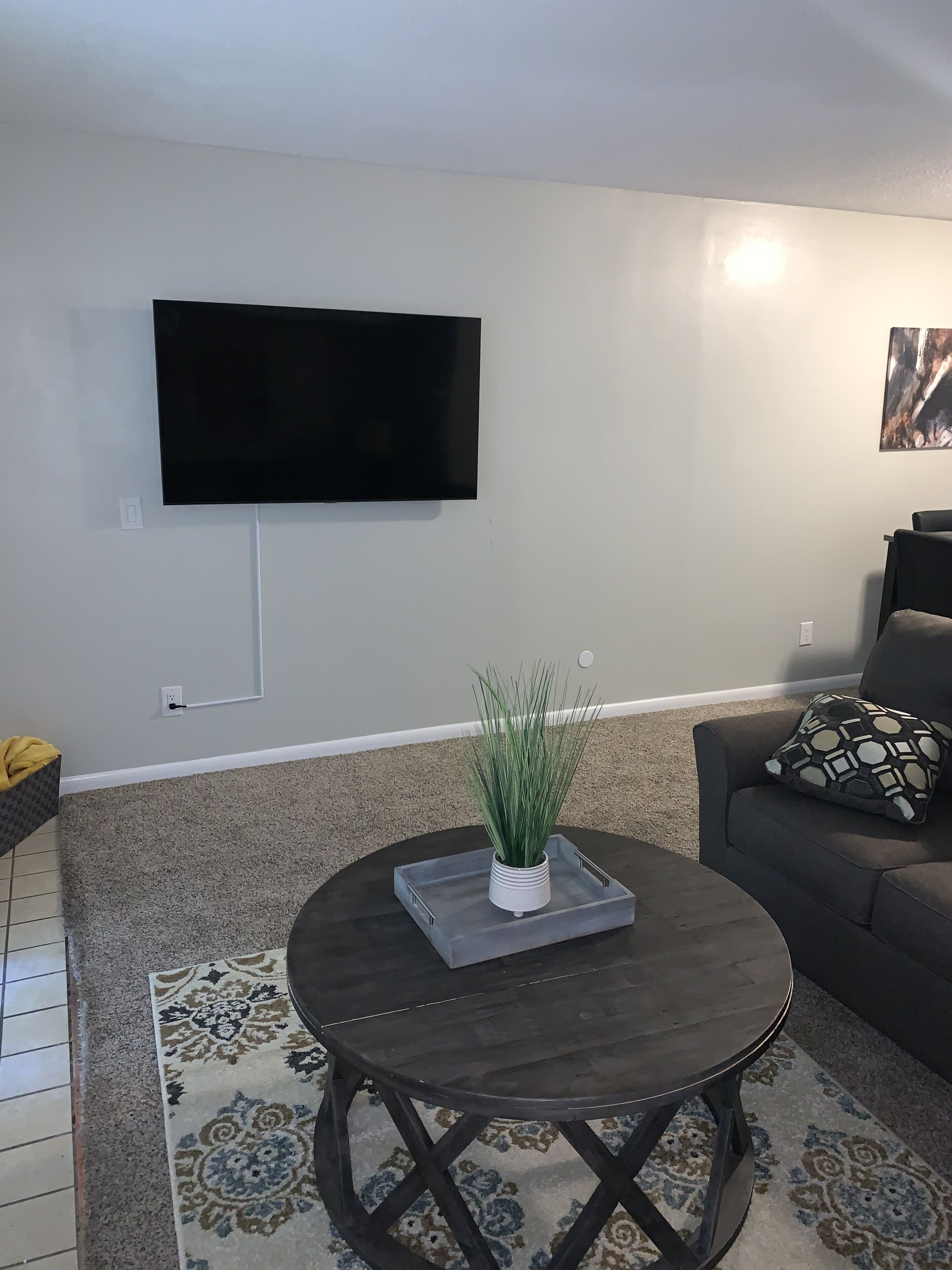 A living room with a flat-screen TV mounted on the wall, a round coffee table with a decorative tray and a potted plant, a dark sofa with patterned cushions, and a patterned rug.