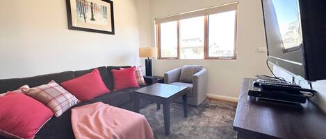 A living room with a black sofa, red and plaid cushions, a grey armchair, a coffee table, a TV on a stand, and a window with blinds.