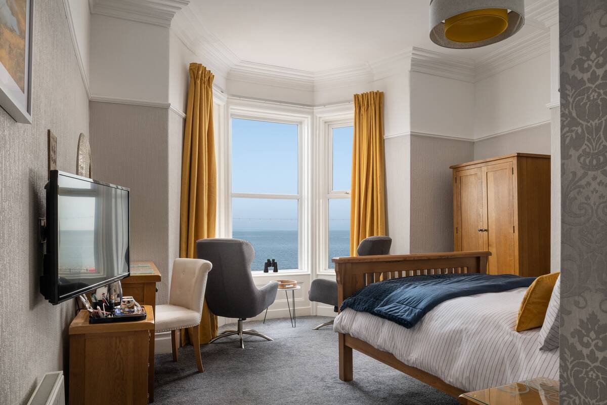 Et hotelværelse med seng, fjernsyn, skrivebord, stol, garderobe og havudsigt gennem store vinduer.