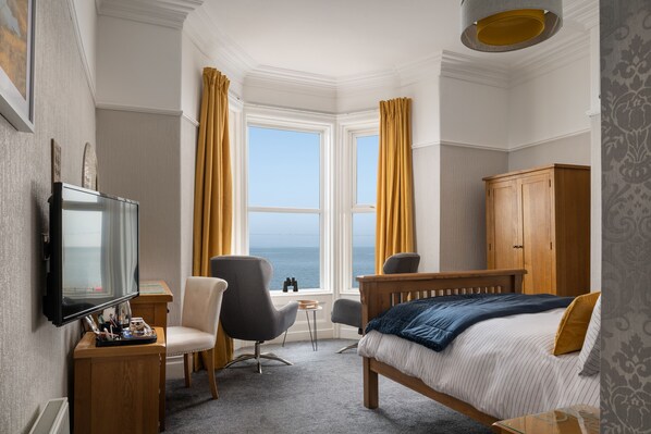 A hotel room with a bed, a TV, a desk, a chair, a wardrobe, and a view of the sea through large windows.