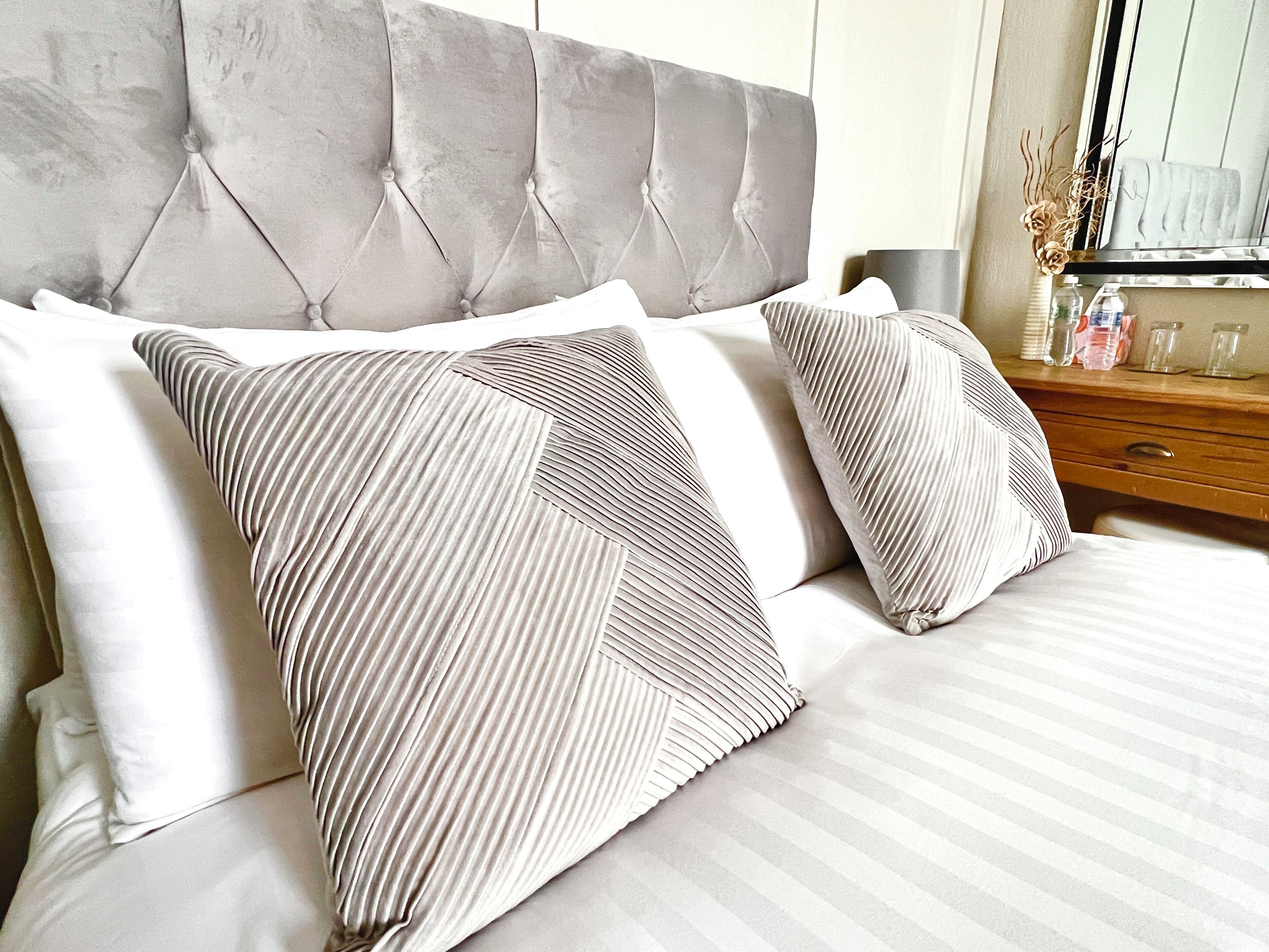 Una cama con cabecera capitonada, dos almohadas con textura de canales y una mesita de noche de madera con un espejo y objetos pequeños.