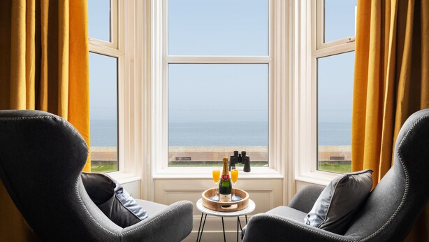 A room with two armchairs facing a large window overlooking the sea, a small table with a bottle and glasses, and binoculars on the windowsill. - The Kendrick Exclusively for Adults (Llandudno)