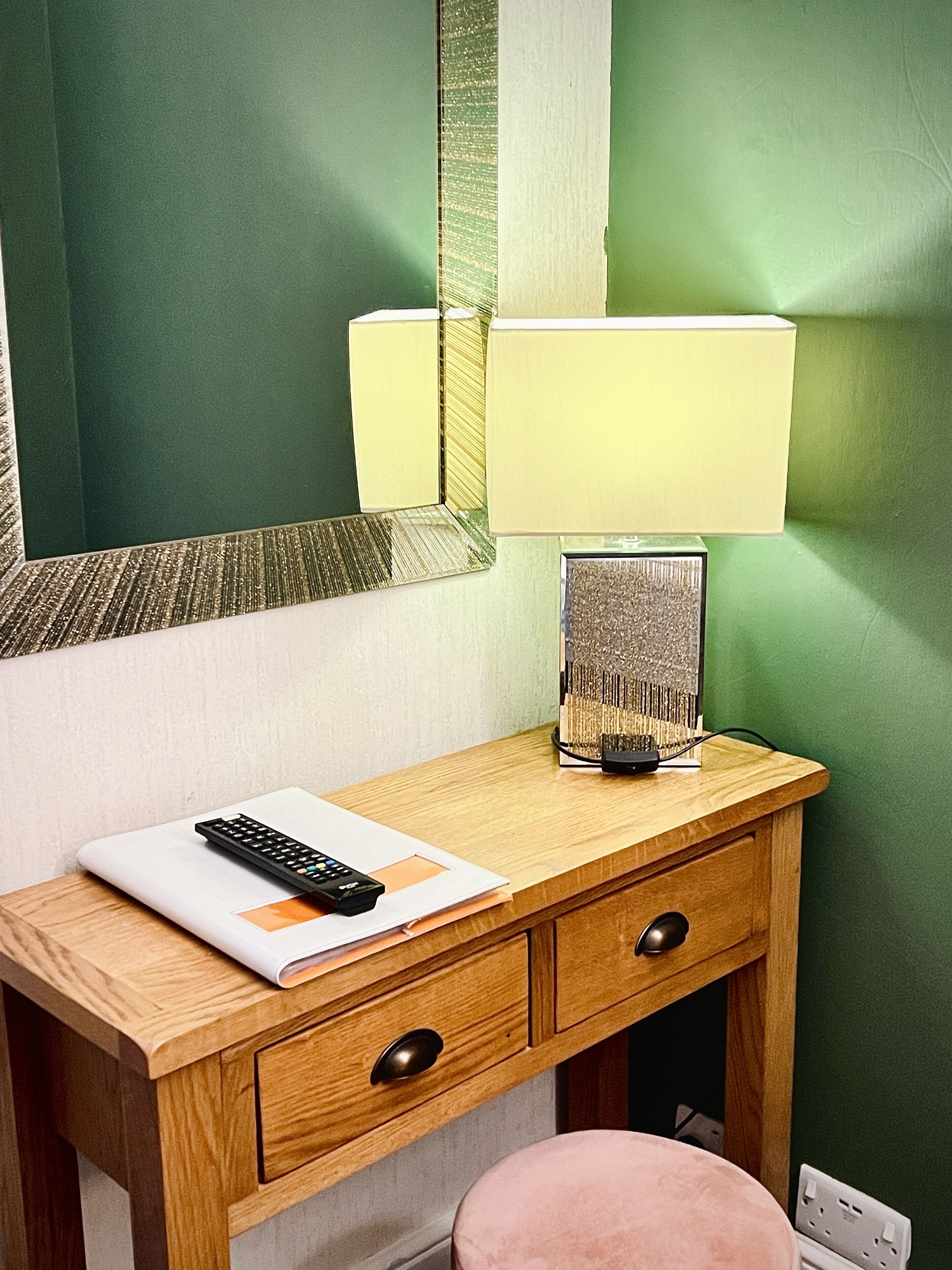 A wooden desk with a lamp, a remote control, and a mirror.