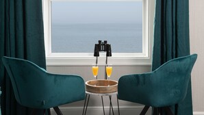 A room with two teal chairs, a small wooden table with binoculars and two glasses of orange juice, and a view of the sea through a window. - The Kendrick Exclusively for Adults (Llandudno)