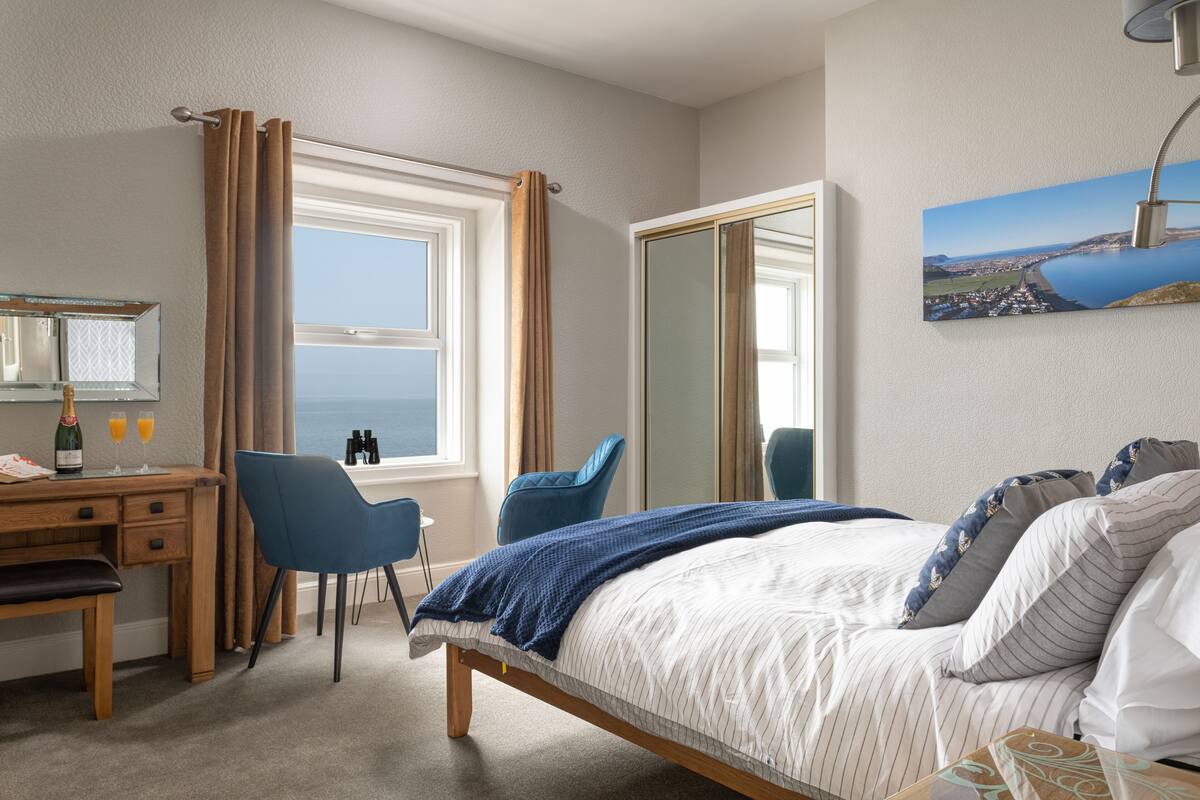 Et hotelværelse med en seng, et skrivebord med stol, et stort vindue med gardiner, et spejl og et maleri af et kystlandskab.