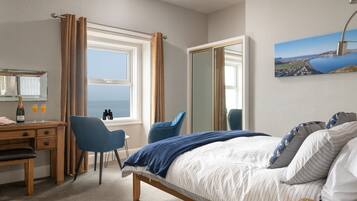 A hotel room with a bed, a desk with a chair, a large window with curtains, a mirror, and a painting of a coastal landscape.