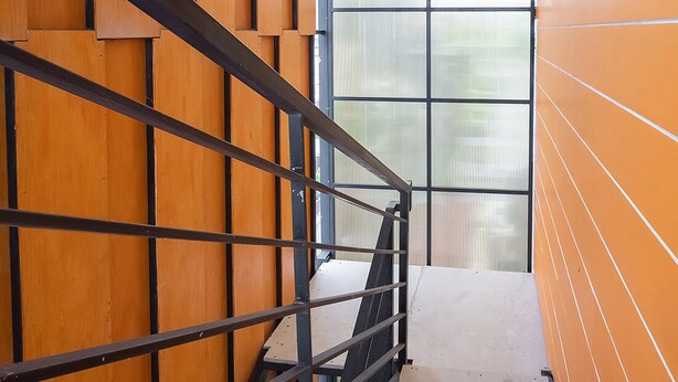 A staircase with metal railings and wooden paneling, leading to a room with a large window. - Graha Asri Syariah (Sidoarjo)