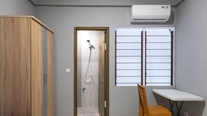 A small room with a wooden wardrobe, a shower area, a window with blinds, a white desk, and a purple bedspread. - Graha Asri Syariah (Sidoarjo)