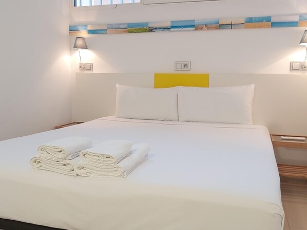 A neatly made bed with white linens and towels, a yellow accent pillow, and two wall-mounted lamps. - Cozy Apartment in Pedregalejo: 300m to Beach, Private Patio, Near Restaurants & Transport (Málaga)