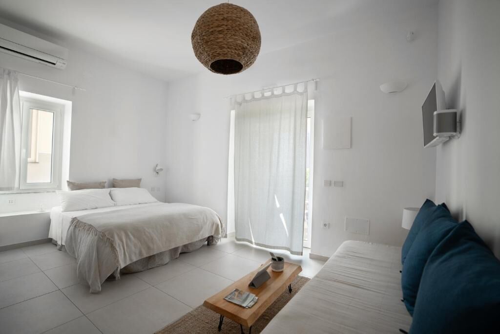 Ein schlicht eingerichtetes Schlafzimmer mit Bett, Sofa, Couchtisch und einem großen Fenster mit Vorhängen.