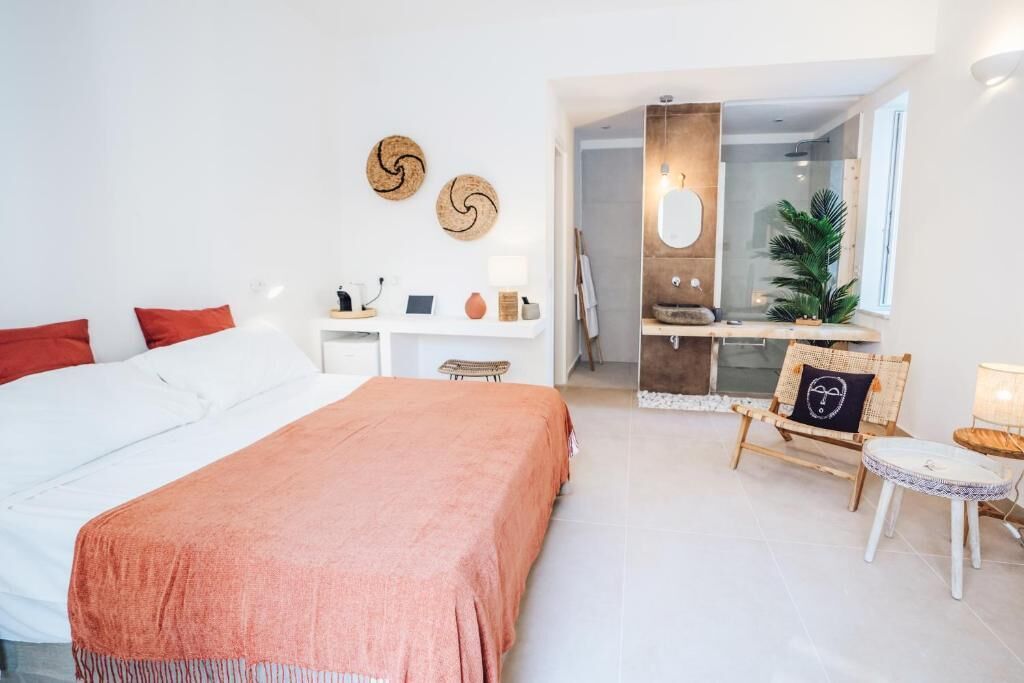 A modern bedroom with a large bed, a bedside table, a small desk, and a chair. There is a bathroom visible through an open door.