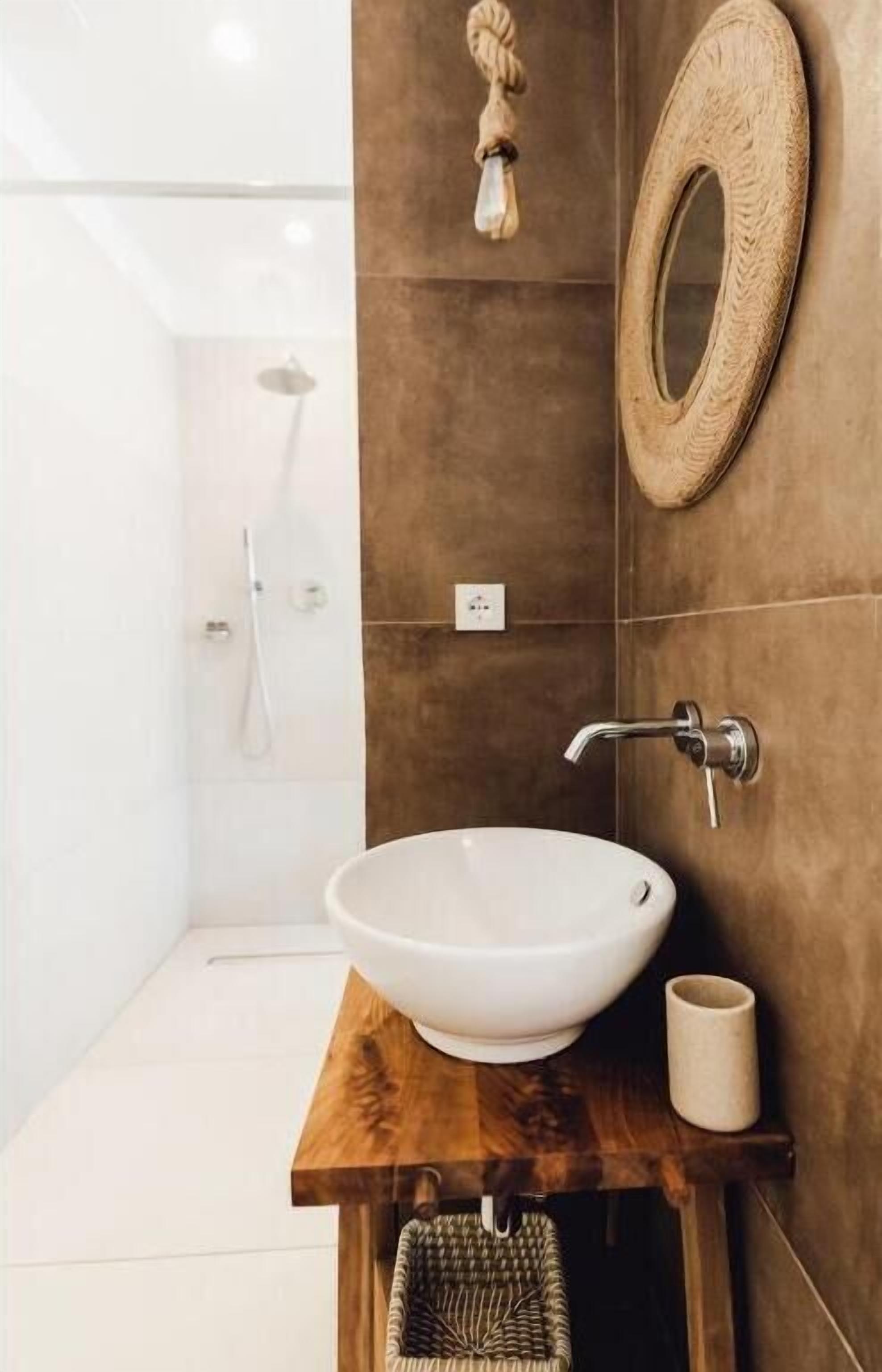 A bathroom with a wooden vanity, a round mirror, a white sink, and a shower area.