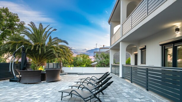 An outdoor seating area with lounge chairs and a table, surrounded by potted plants and palm trees, with a building in the background. - Villa LiLi Apt 2 with balcony (Poreč)