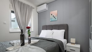 A modern bedroom with a grey bed, white pillows, a bedside table with a lamp, a window with curtains, and a wall-mounted air conditioner. - Villa LiLi Apt 2 with balcony (Poreč)