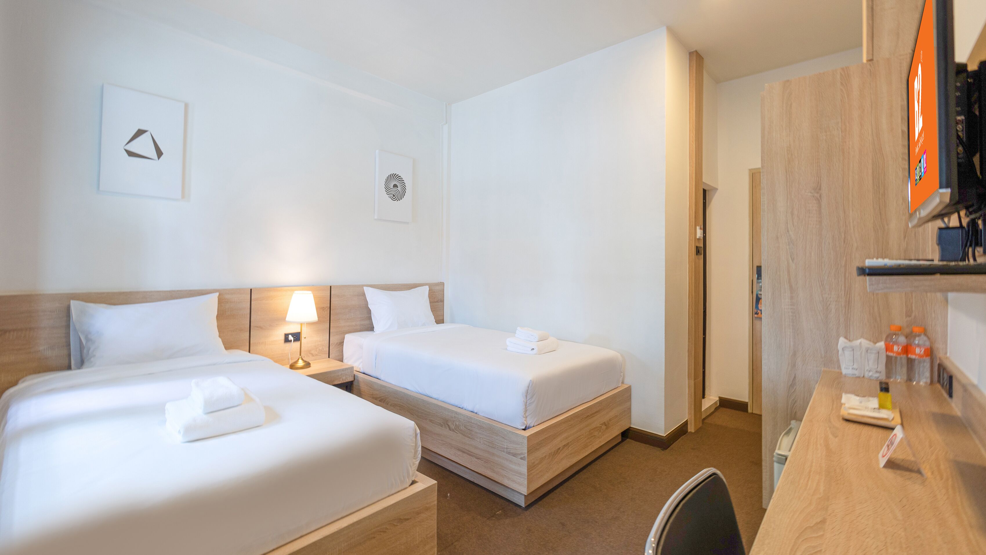 Habitación de hotel con dos camas individuales, un escritorio con silla, una lámpara y un estante pequeño con artículos de aseo.
