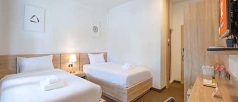 Habitación de hotel con dos camas individuales, un escritorio con silla, una lámpara y un estante pequeño con artículos de aseo.