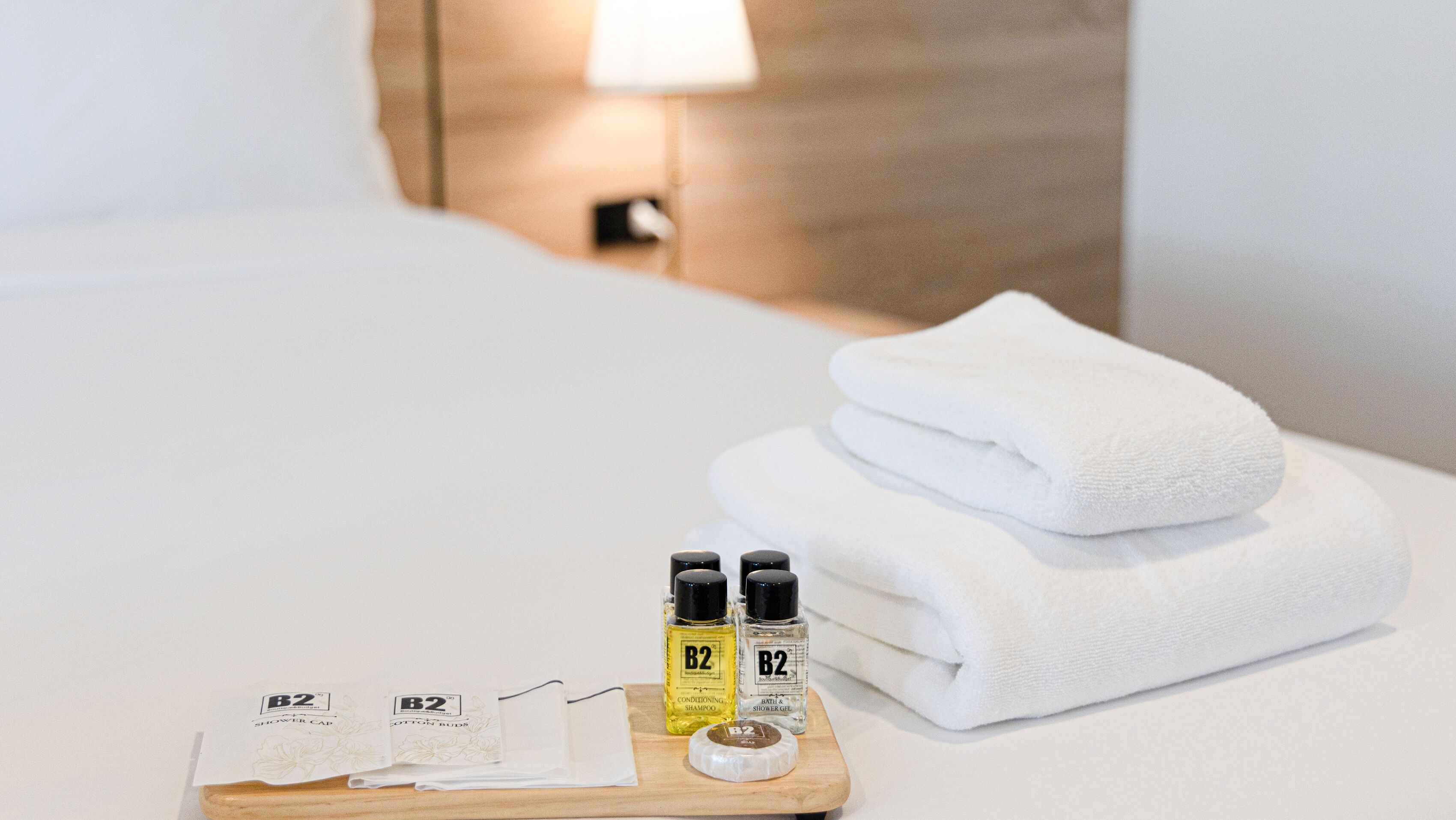 A neatly made bed with a folded white towel and a tray with toiletries including B2 conditioning shampoo, B2 shower gel, and B2 body spray.