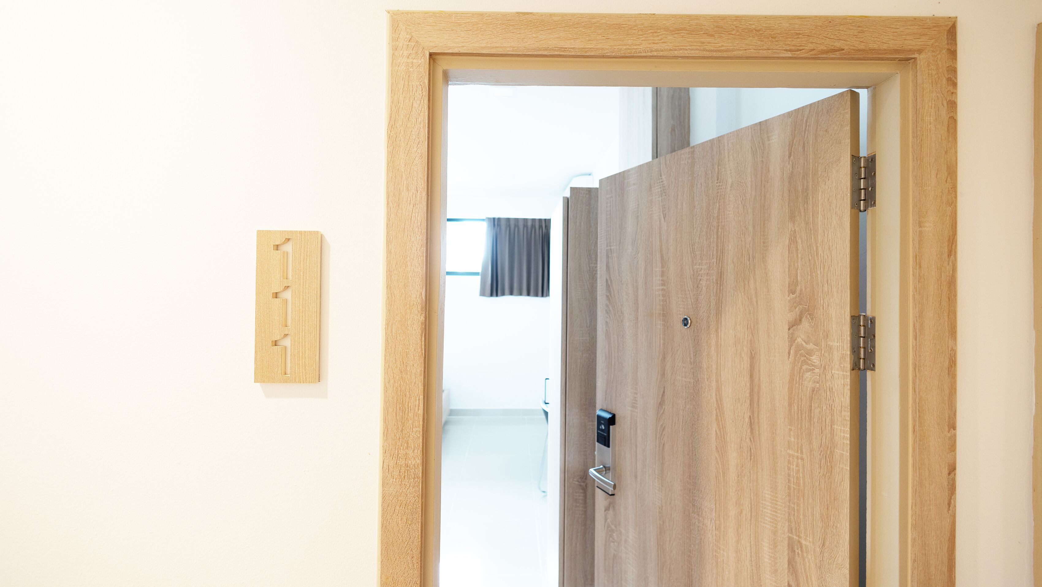 A wooden door with a metal handle and a latch, leading to a room with a white wall and a window with a curtain.