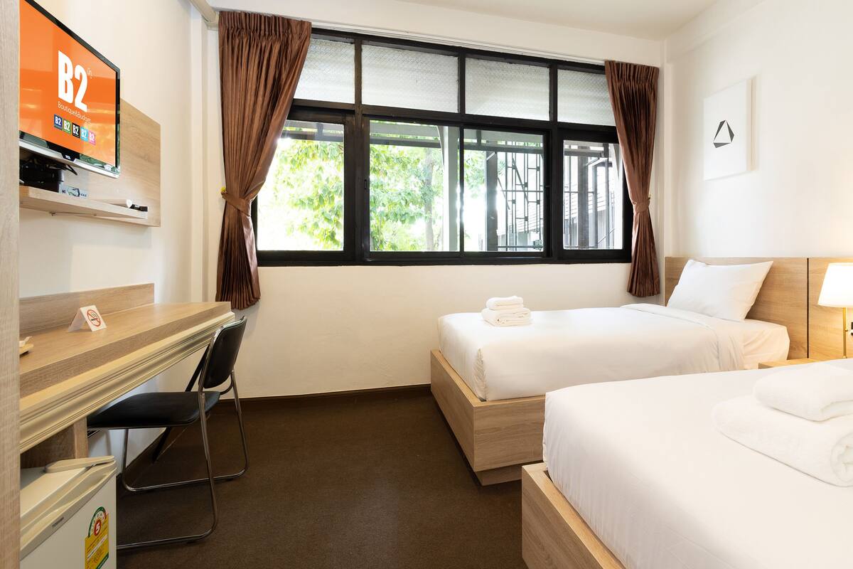Habitació d'hotel amb dos llits, un escriptori, una cadira, un televisor i una finestra amb cortines.