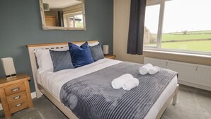 A neatly made bed with a grey and white striped blanket, two pillows, and two rolled towels on the bed. There is a wooden nightstand with a lamp and a drawer, a mirror on the wall, and a window with a view of a green field. - Coedfan (Ruthin)