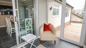 A modern living room with a sliding glass door leading to a patio, a dining area with a glass table and chairs, and a small white table. - Coedfan (Ruthin)