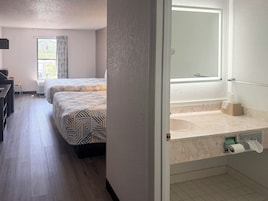 Habitación de hotel con dos camas, un escritorio, una ventana con cortinas y un baño con espejo y dispensador de toallas.