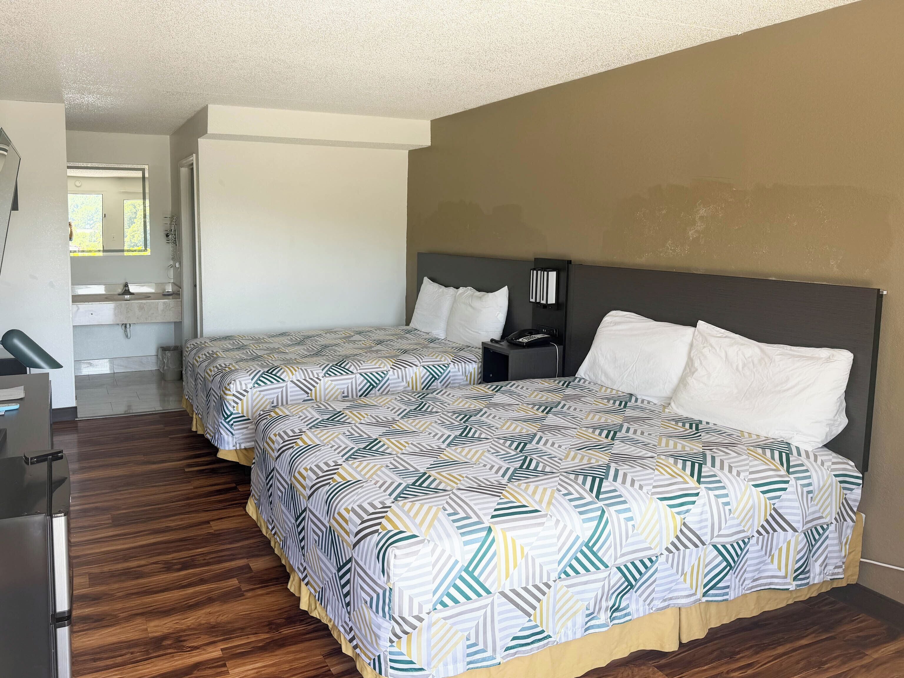 Habitación de hotel con dos camas, una mesita de noche, un escritorio y baño con espejo y lavamanos.