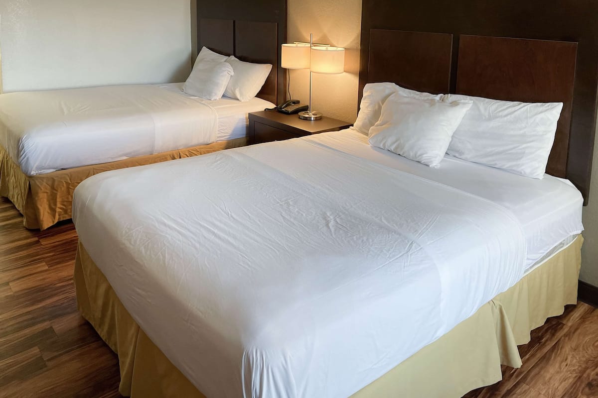 Habitación de hotel con dos camas, cada una con ropa de cama blanca y cabeceros de madera. Hay una mesita de noche con una lámpara y un teléfono. El piso es de madera y las cortinas son color beige.