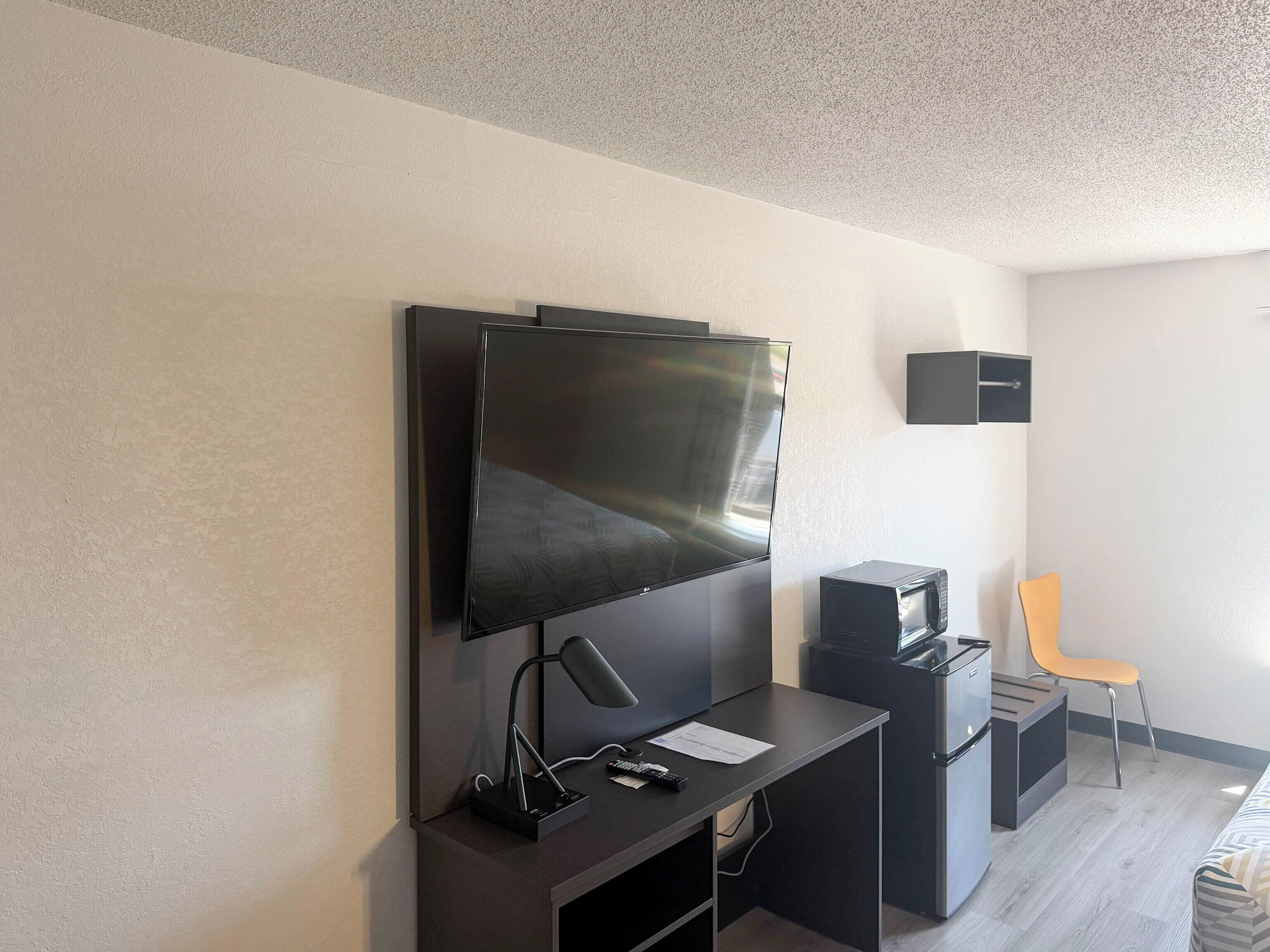 A modern room with a flat-screen TV mounted on the wall, a desk with a lamp, a microwave, and a chair.