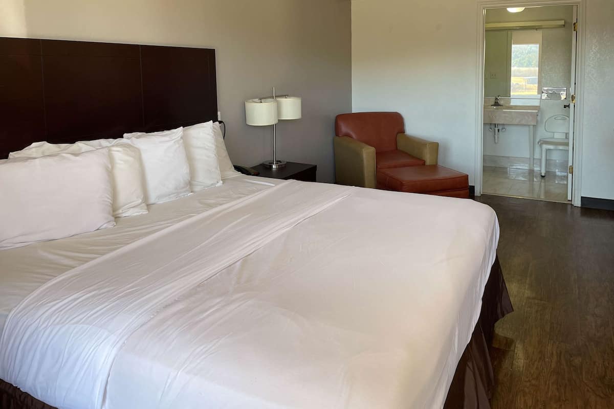 Habitación de hotel con una cama grande, una mesita de noche con lámparas, un sillón de cuero y vistas al baño a través de una puerta abierta.