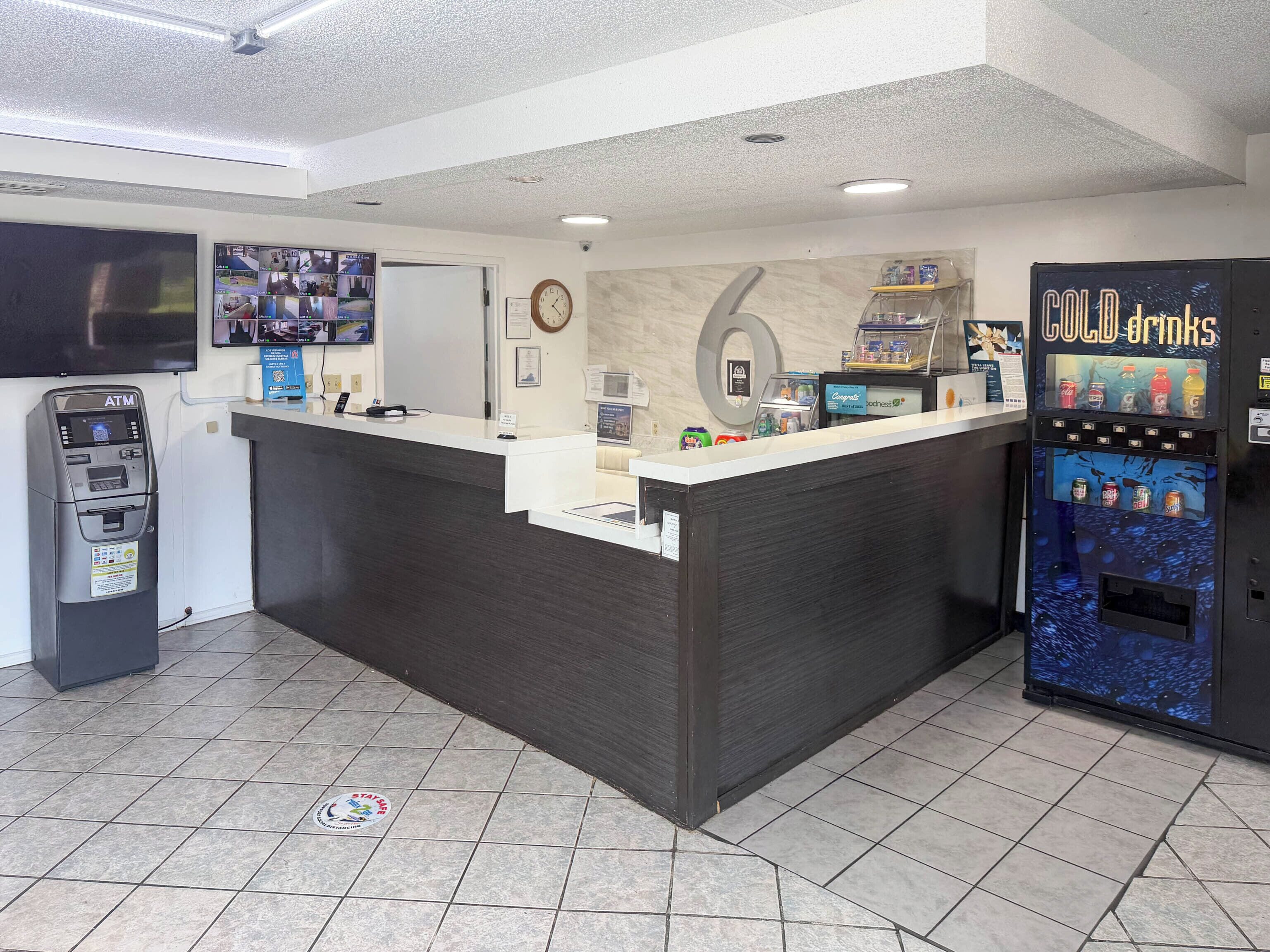 A reception area with a counter, an ATM, a CCTV monitor, a clock, and a vending machine labeled 'COLD DRINKS'.