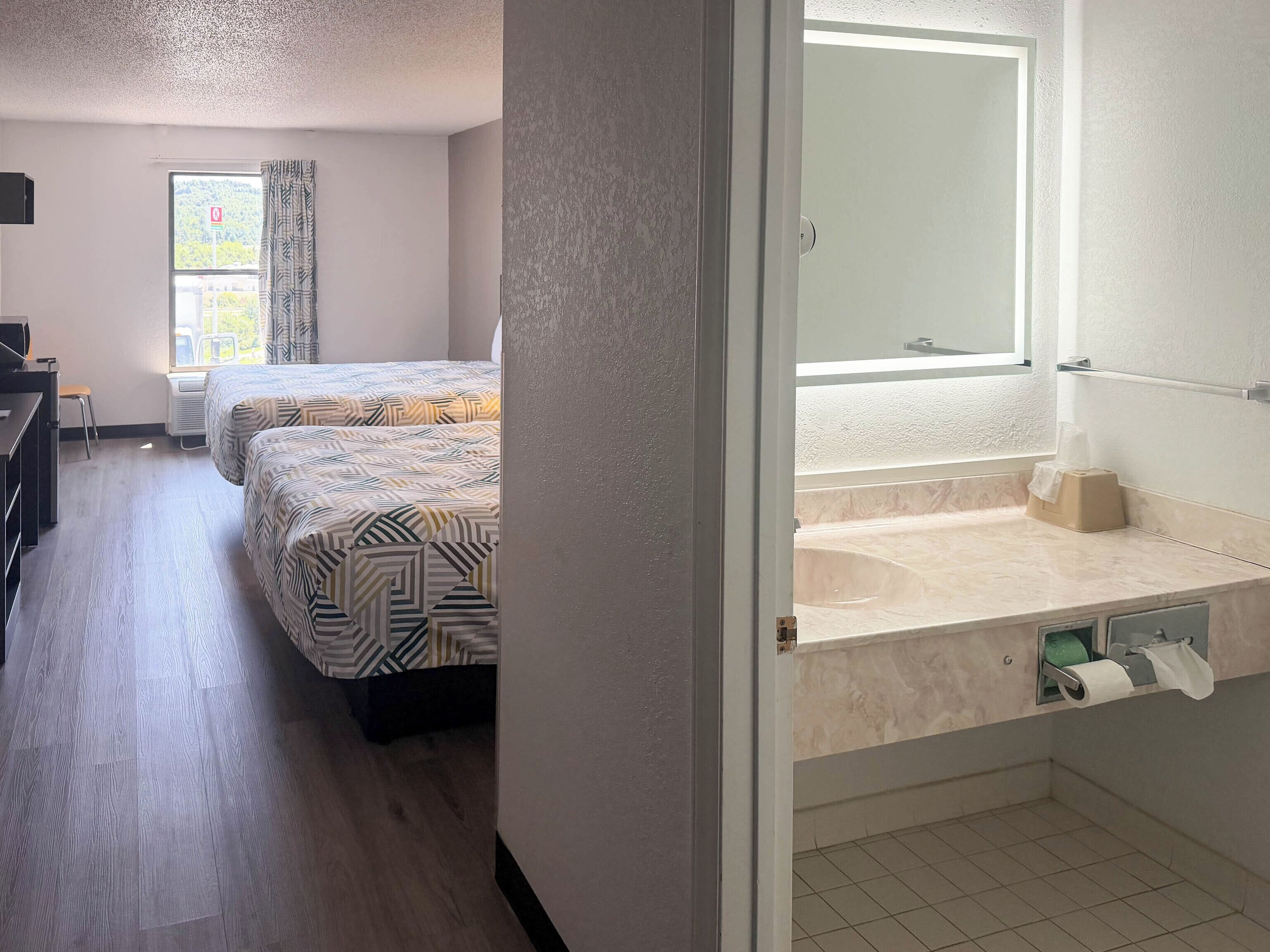 A hotel room with two beds, a desk, a chair, a window with curtains, and a bathroom with a sink and toilet.
