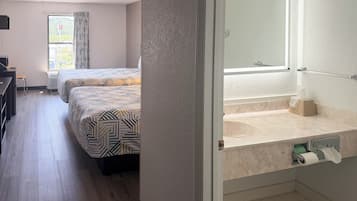 A hotel room with two beds, a desk, a chair, a window with curtains, and a bathroom with a sink and toilet.