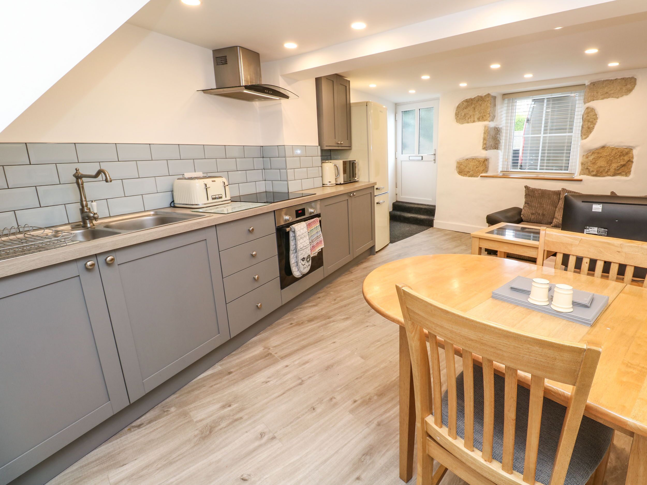A modern kitchen with grey cabinets, a stainless steel range hood, a built-in oven, and a dining area with a wooden table and chairs.