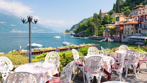 Une terrasse en bord de mer, agrémentée de chaises et de tables blanches décorées, qui offre une vue sur un port où se trouvent des bateaux et des bâtiments aux couleurs vives.
