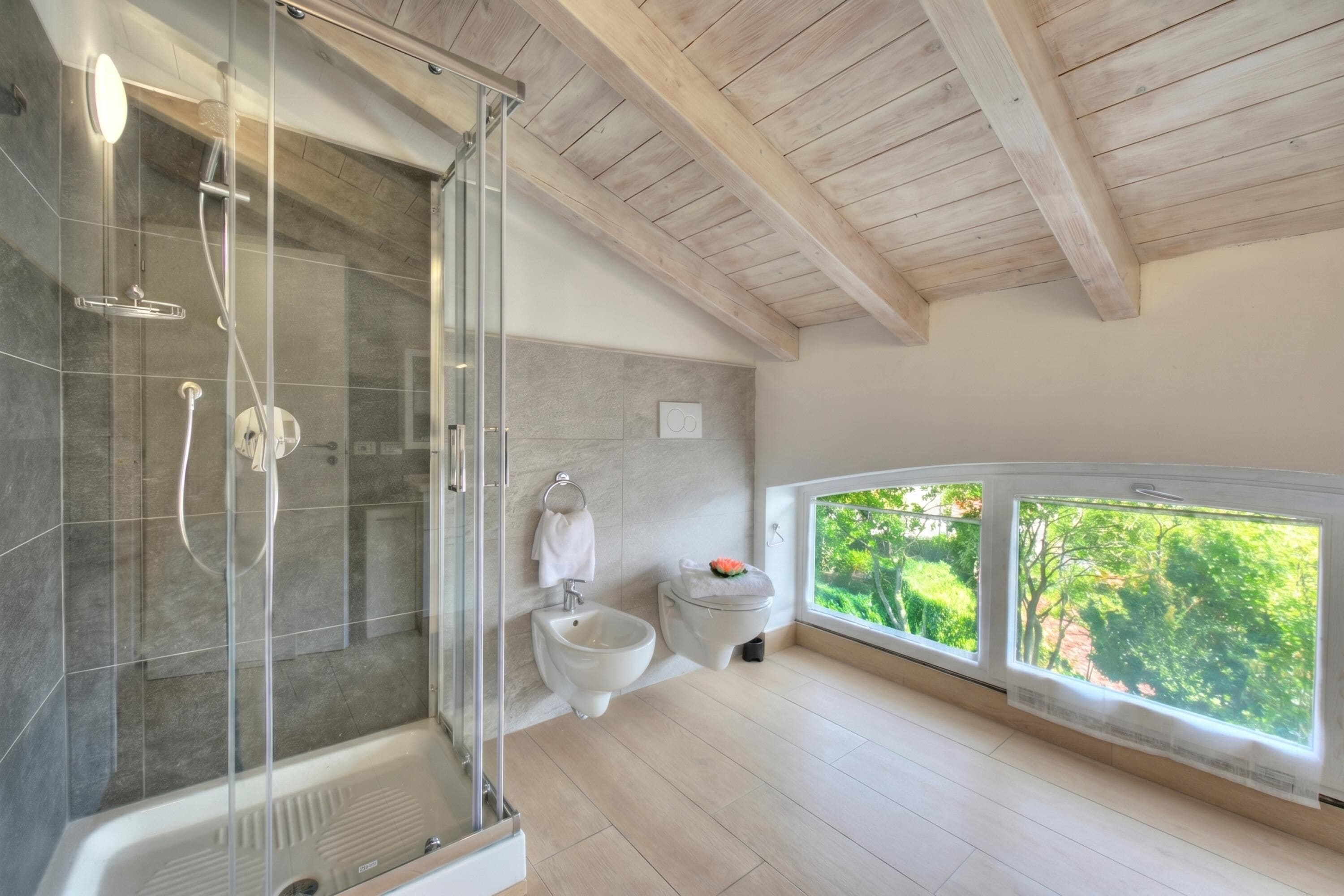 A modern bathroom with a glass-enclosed shower, a bidet, and a large window offering a view of greenery.