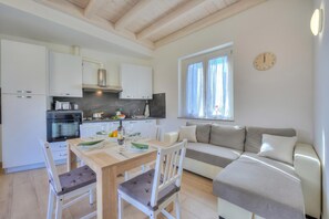 A modern kitchen with white cabinets, a dining table with chairs, and a sofa in a living area with a clock on the wall.