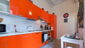 Ett kök med orange skåp, en mikrovågsugn, ett handfat och ett litet bord med en vattenkokare och koppar.
