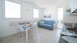 A modern kitchen with white cabinetry, a dining area with a table and chairs, a blue sofa, and a large window providing natural light. - Stylish Residenza Torre Bianca 2 Bedroom Sleeps 6 (Lignano Sabbiadoro)
