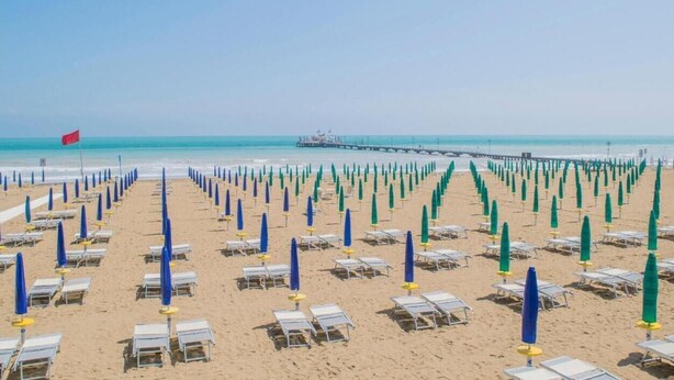 A beach with rows of sun loungers and umbrellas, a pier in the background, and a flag on the left. - Stylish Residenza Torre Bianca 2 Bedroom Sleeps 6 (Lignano Sabbiadoro)