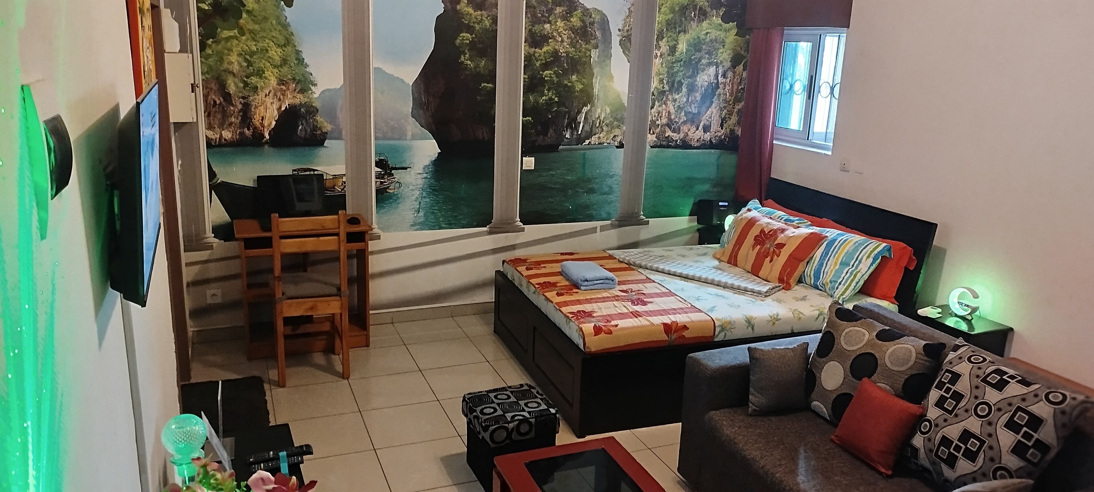 A bedroom with a bed, a desk, a chair, a sofa, and a view of a coastal landscape through large windows.