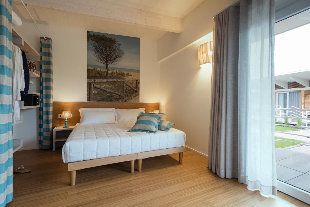 A bedroom with a wooden bed, a nightstand with a lamp, a painting on the wall, and a view of the outside through a large window.