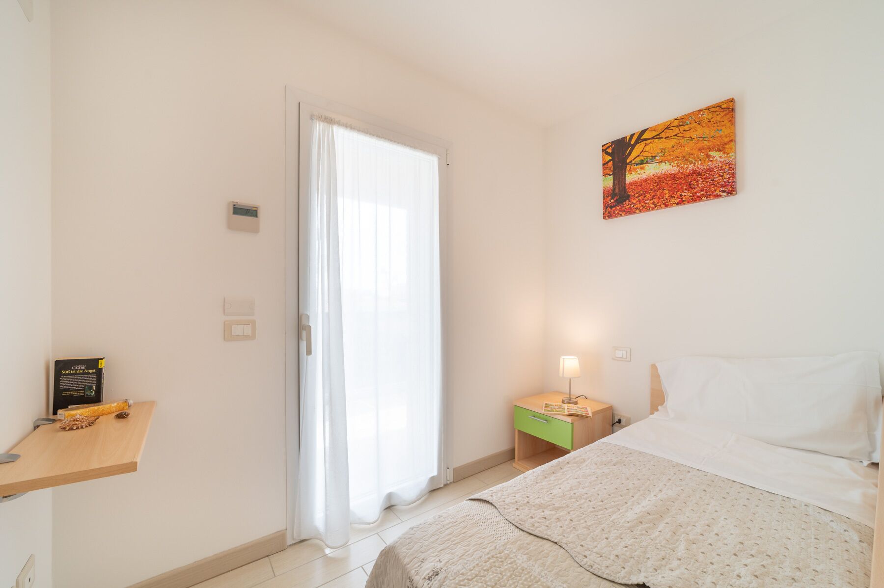 Ein Schlafzimmer mit einem Bett, einem Nachttisch, einer Lampe, einem Gemälde, einem Fenster mit Vorhängen und einem Thermostat.