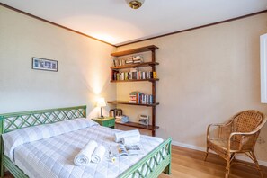 Una cama bien hecha con cabecera verde, una estantería de madera, un sillón de mimbre y una mesita de noche con lámpara.
