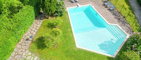 A rectangular swimming pool with a stone border, surrounded by a stone pathway, and a grassy area with a few trees and bushes.