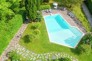 Una piscina rectangular con borde de piedra, rodeada por un camino de piedra y un césped con algunos árboles y arbustos.
