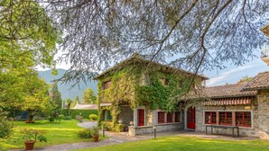 A stone building with red doors and windows, surrounded by a well-maintained garden and trees. - Villa Colle (Tremezzina)