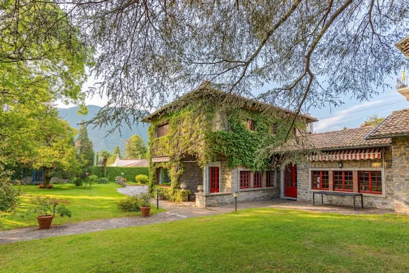 A stone building with red doors and windows, surrounded by a well-maintained garden and trees. - Villa Colle (Tremezzina)