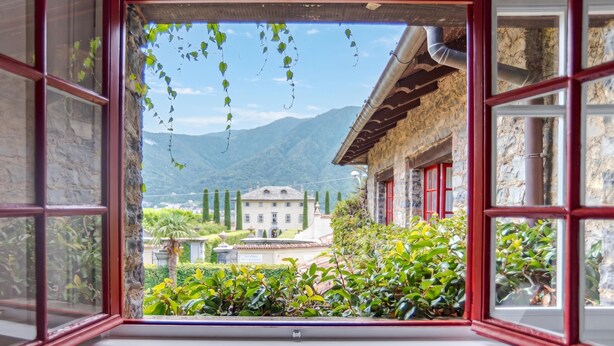 A view through an open window with a stone building, greenery, and mountains in the background. - Villa Colle (Tremezzina)