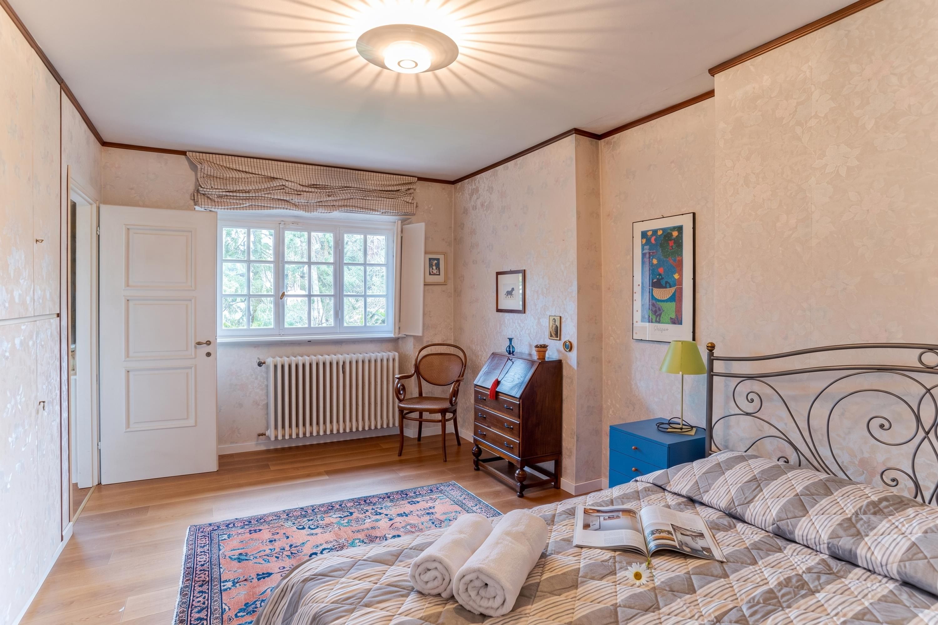 Ein Schlafzimmer mit Bett, Kommode, Stuhl, Heizkörper, Fenster und einem Bild an der Wand.